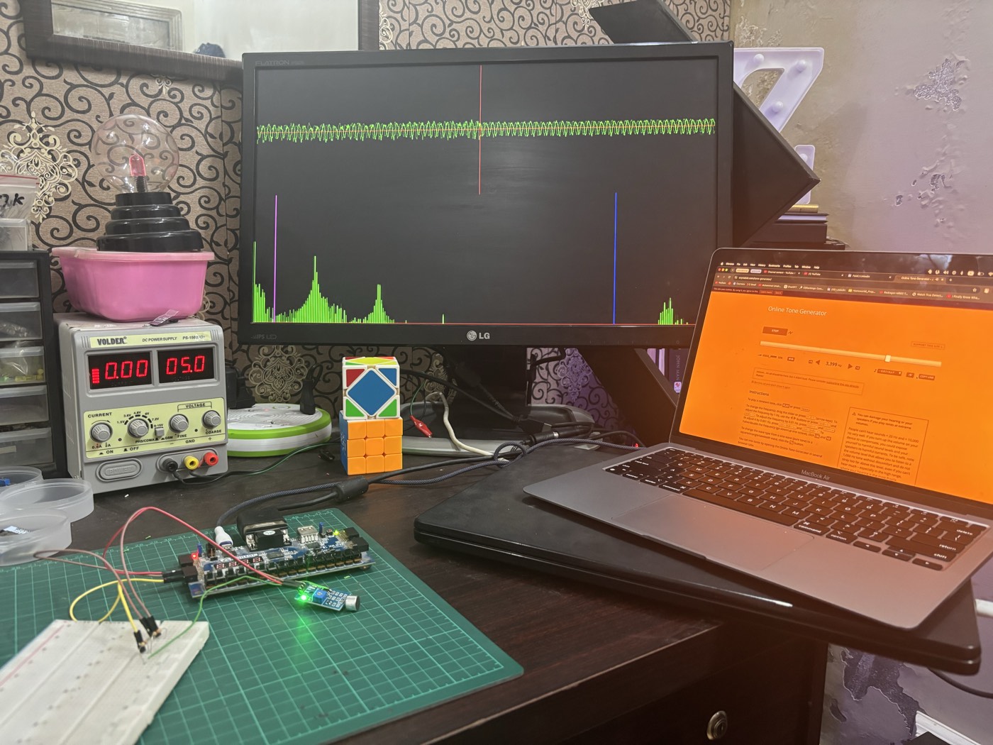 The full bench setup: an LG monitor showing the live waveform and 256-bin spectrum with pink and blue cutoff markers, the Basys3 board wired to the sound sensor on a breadboard, and a laptop running a tone generator for testing